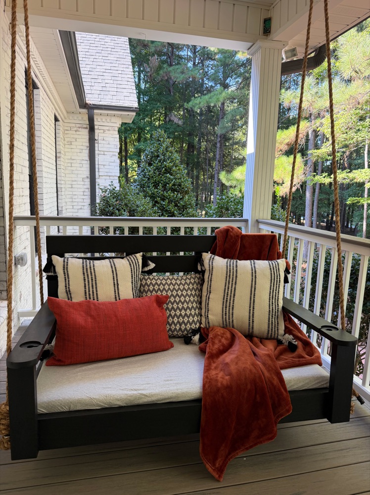 Black-framed bed swing with a rust throw blanket and patterned pillows on a covered porch